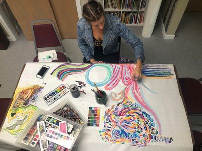 Allison at a table covered in watercolors and pastels, drawing sweeping rainbow swirls on a long sheet of paper.