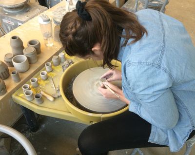 Allison at a pottery wheel, shaping a small clay cup, with a row of freshly thrown cups on the table beside her.